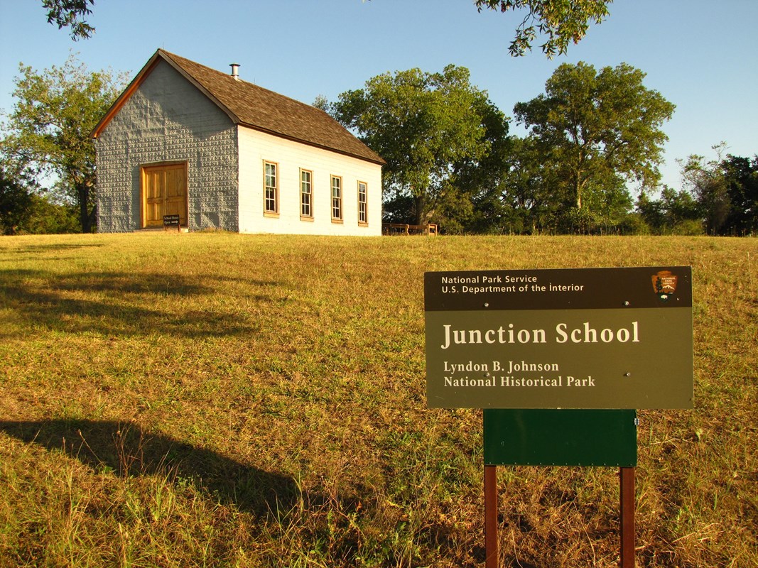 Small, gray building on a hill. Sign in front reads