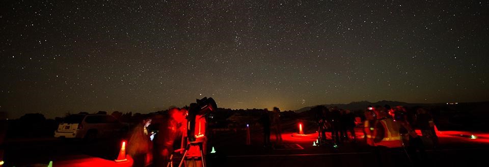 people gather around telescopes illuminated red under a starry sky