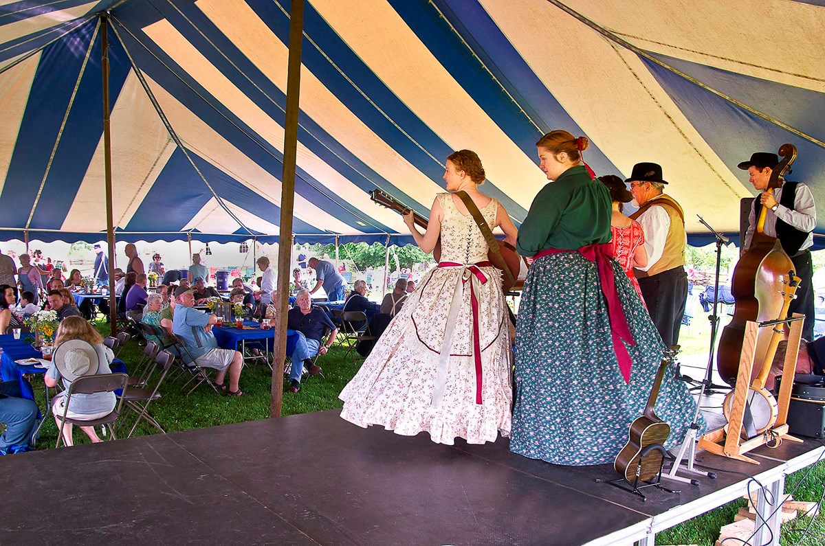Band in Civil War period clothing playing on a stage in front of audience sitting under a tent