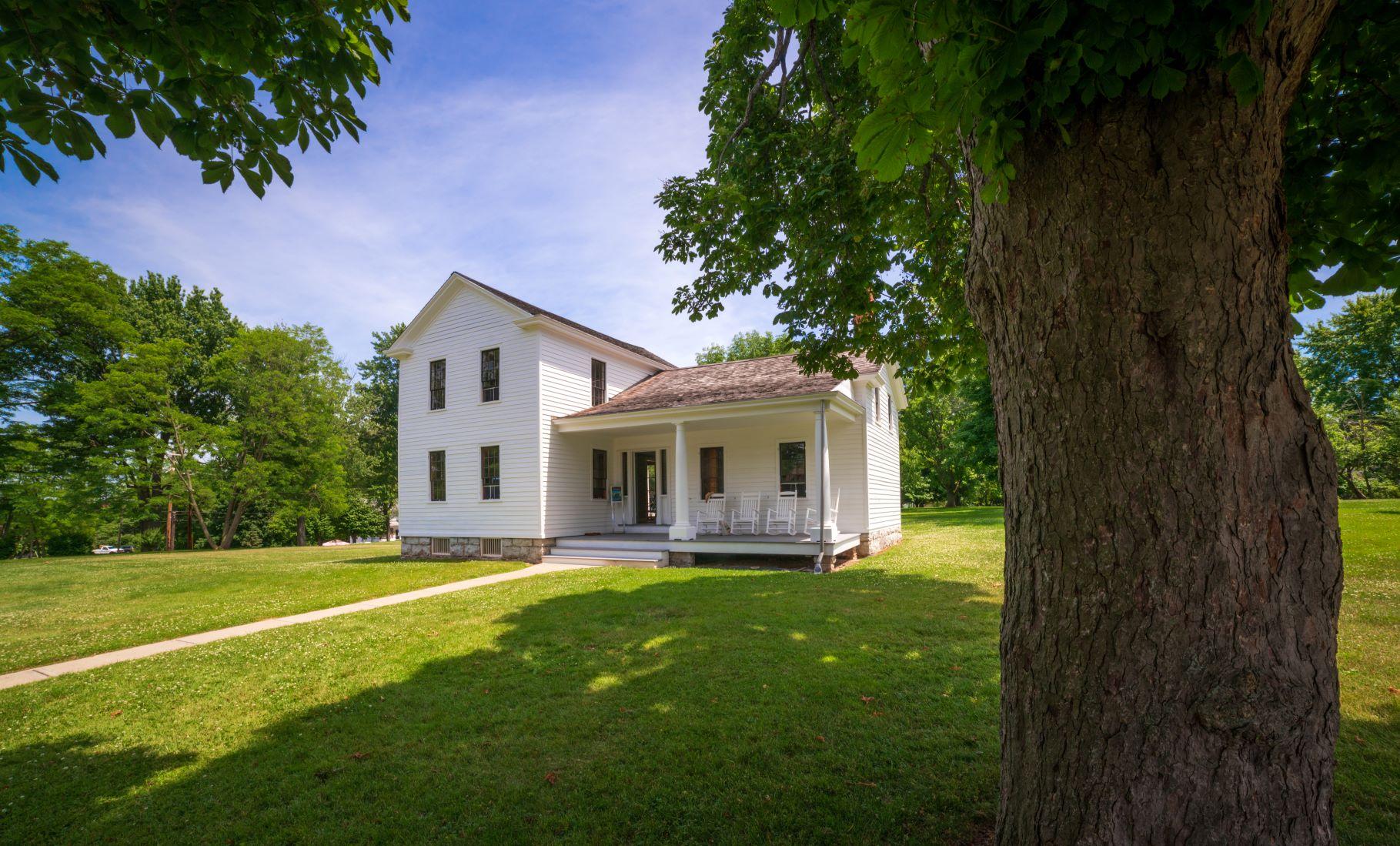 A white clapboard house. One and a half stories tall, surrounded by green lawn and trees.