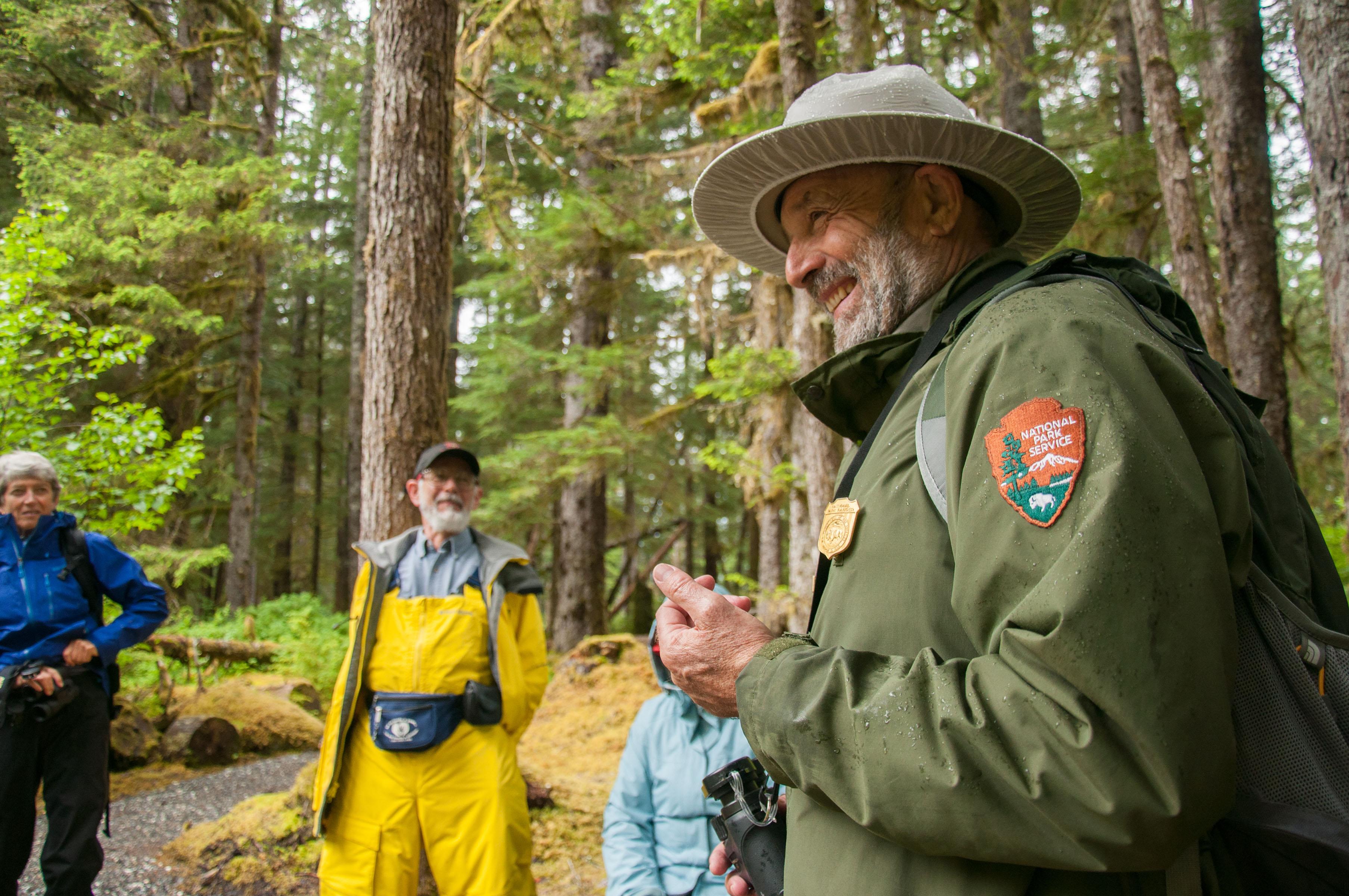 Park Ranger smiles on a forested path with visitors onlooking.