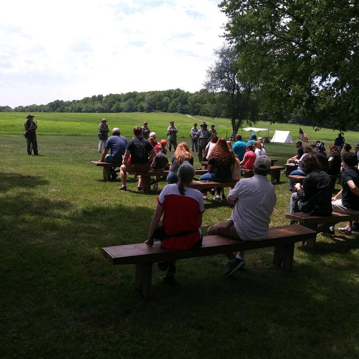 An audience sits on benches watching reenactors and living historians during a talk on a sunny day.