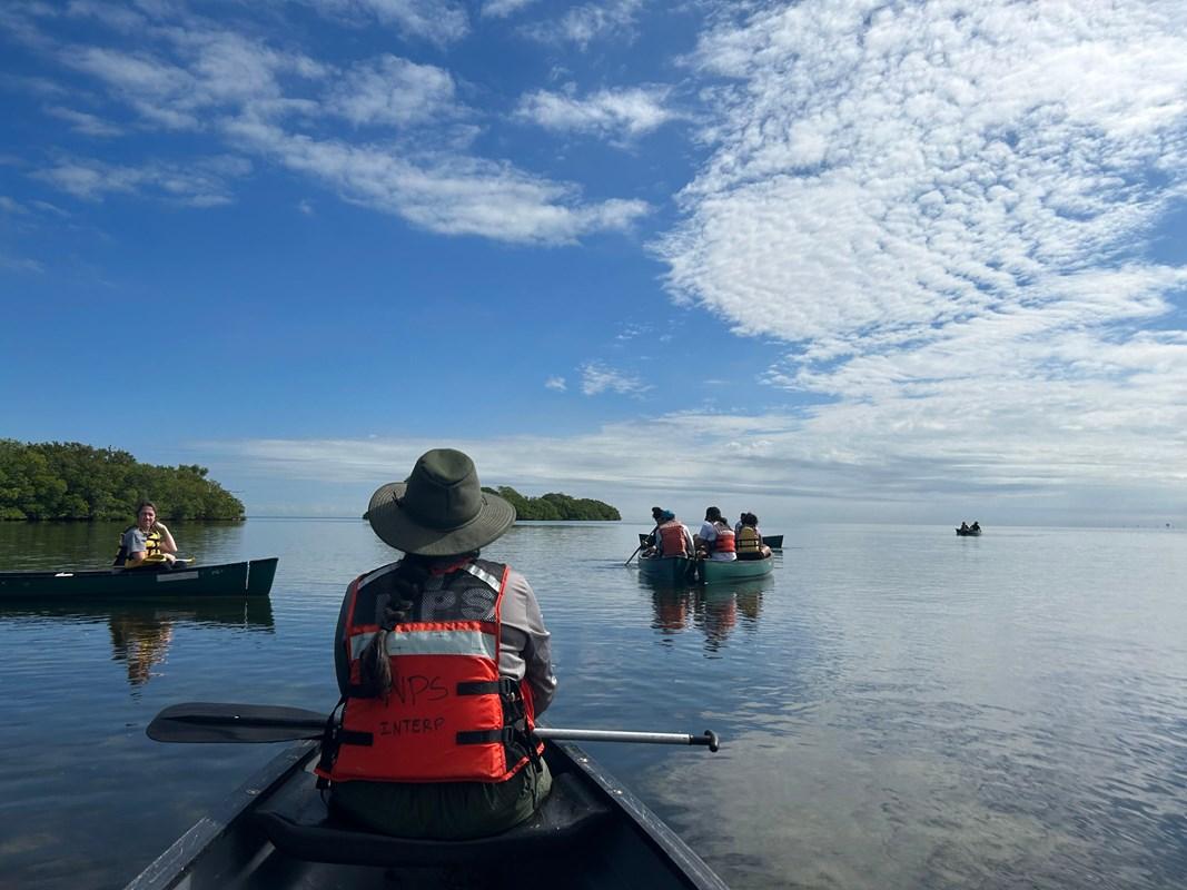 A person sits in the front of a canoe facing away, other canoes are visible on calm waters.