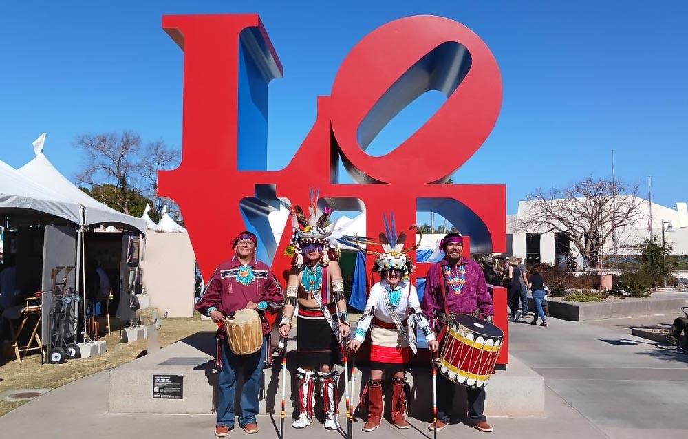 Four Native American dancers in ceremonial clothes stand in front of the word "Love".
