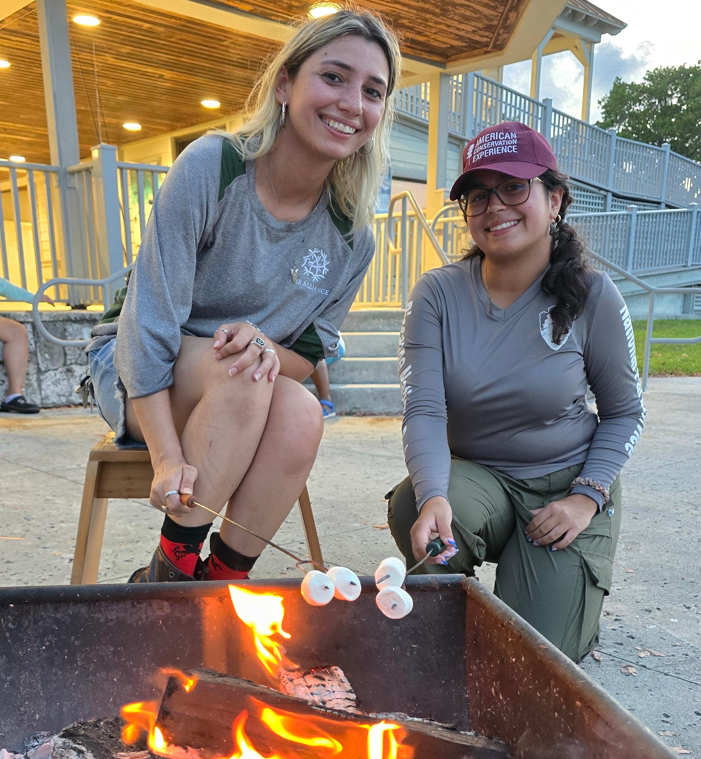 Two smiling young women roast marshmallows over a firepit in front of the visitor center.