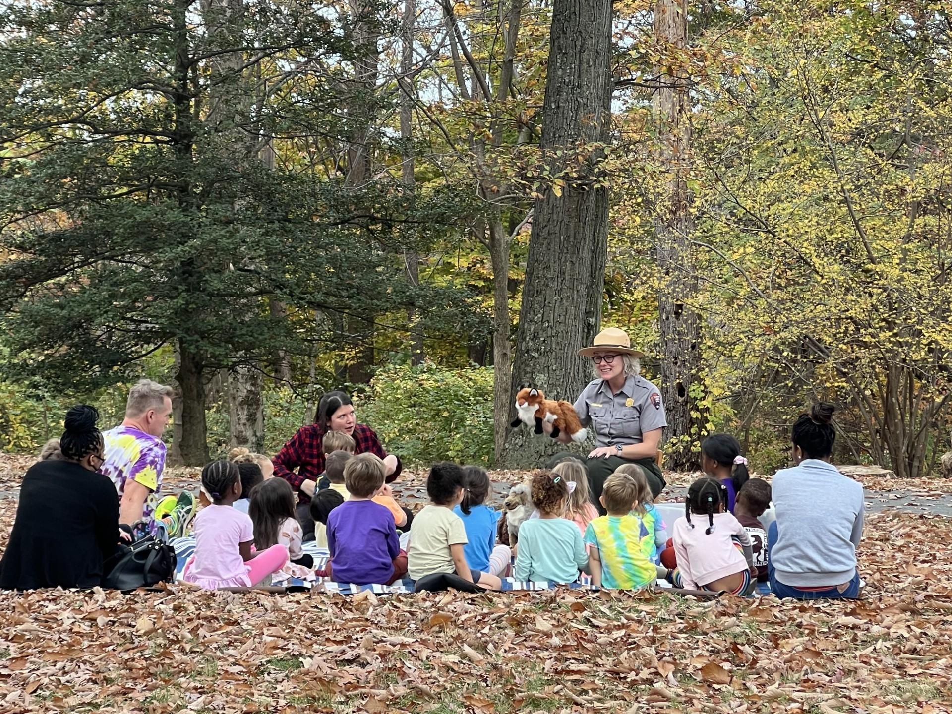 a group of young children sit on the forest floor listening to a park ranger with a fox hand puppet