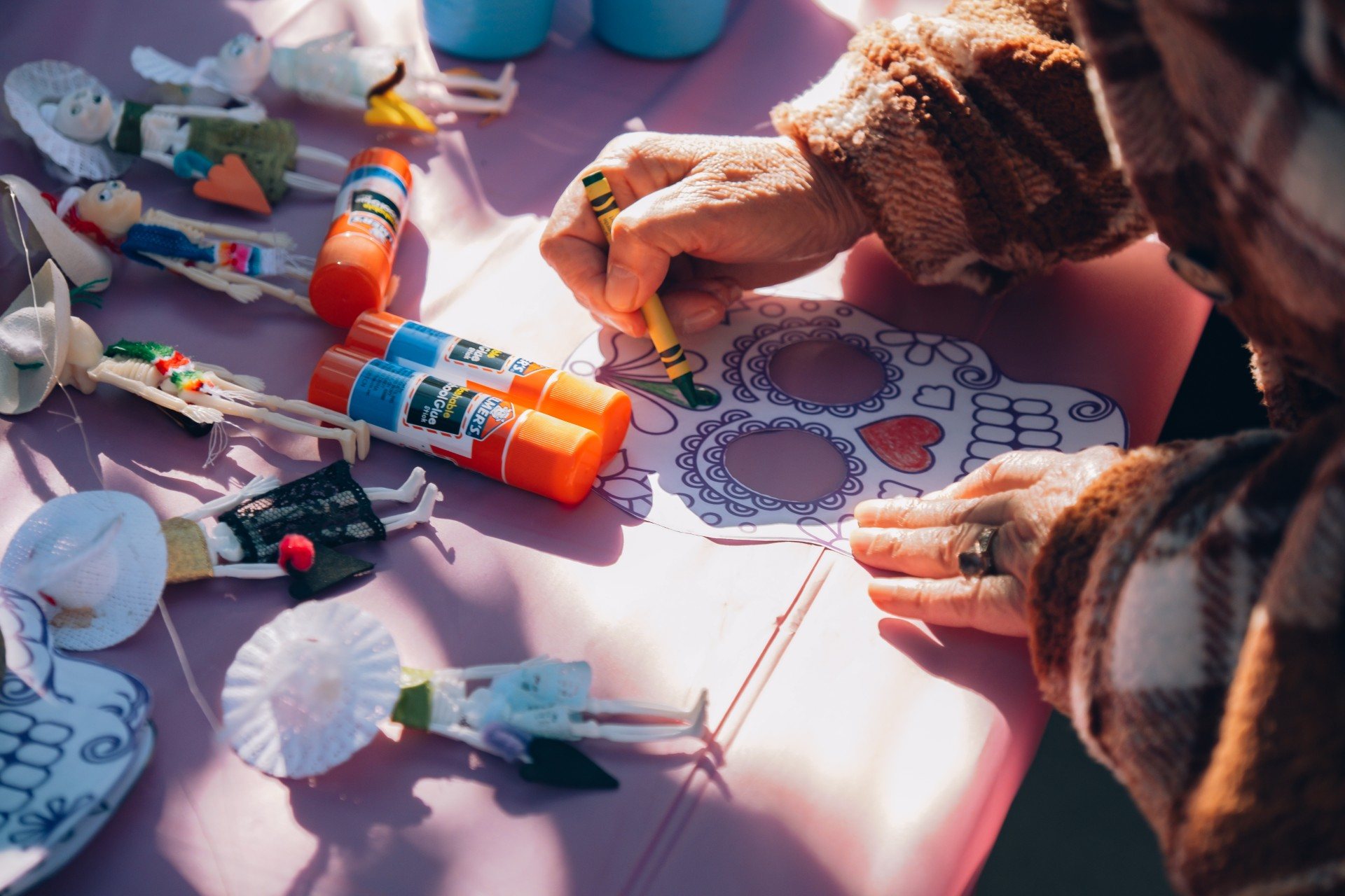 table top covered in craft supplies and art; an older person's hands are seen coloring a paper skull