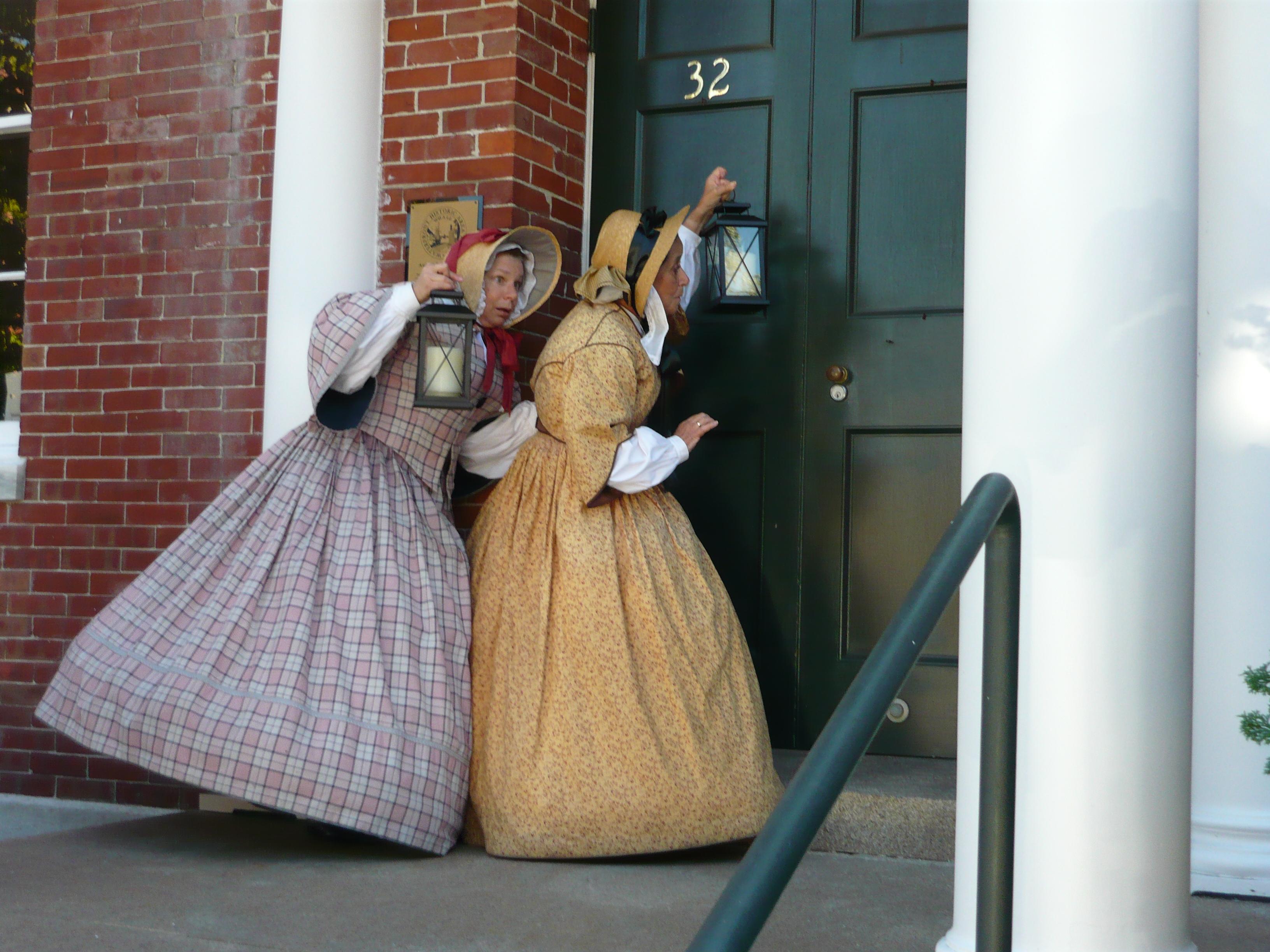 Two women wearing dresses from the 1850s and carrying lanterns knock on the door of an historic buil