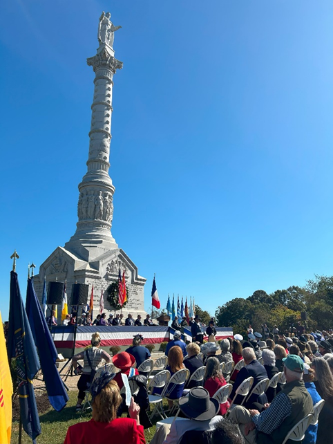 A crowd of people surrounding a stage with blue sky and Yorktown Victory Monument.