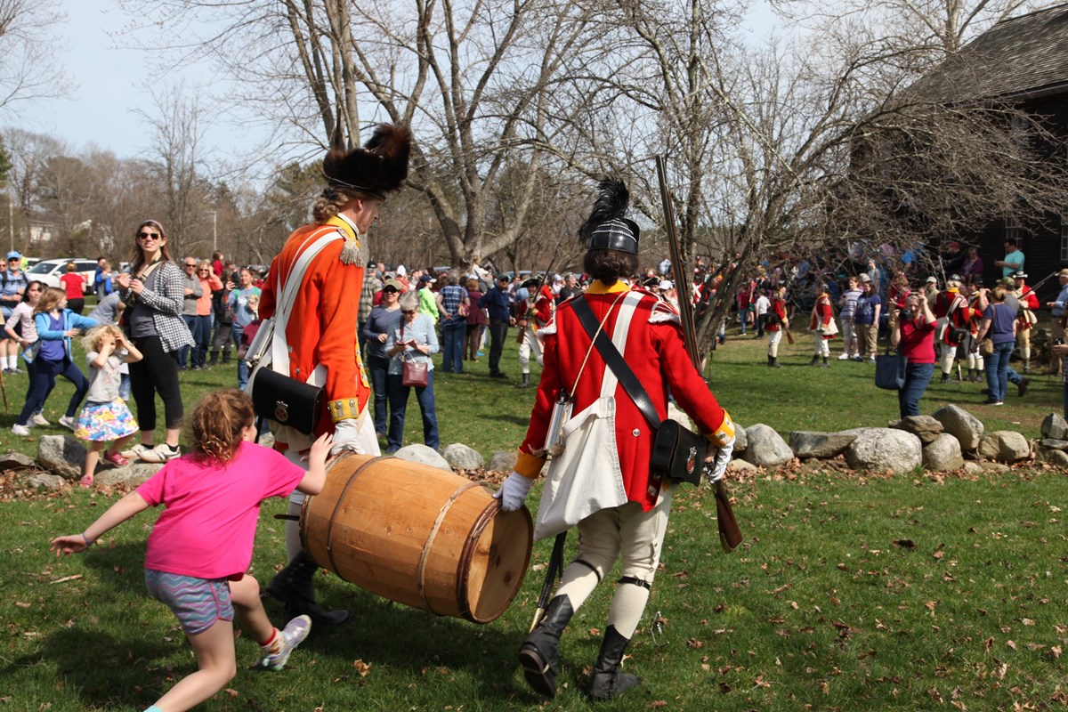 Two red-coat British soldiers carry off a wooden barrel while child runs behind them.