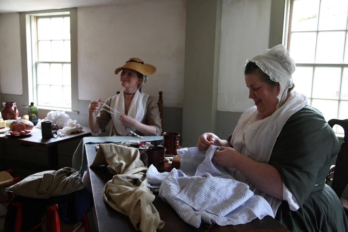Two colonial women sit in a kitchen preparing food and clothing for soldiers