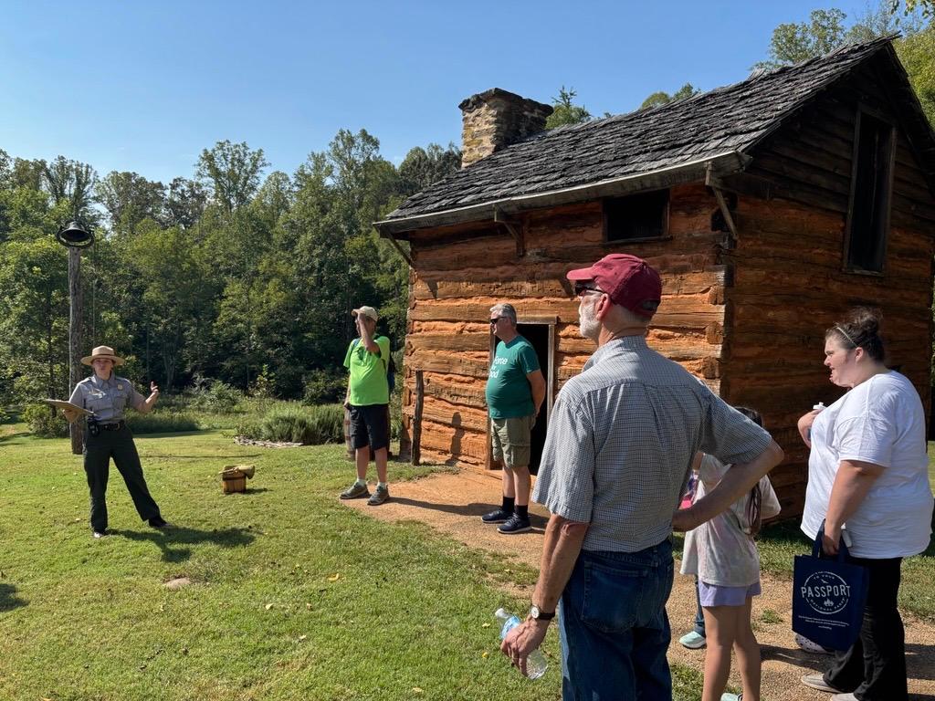 A park ranger speaks to visitors in front of a brown wooden cabin.