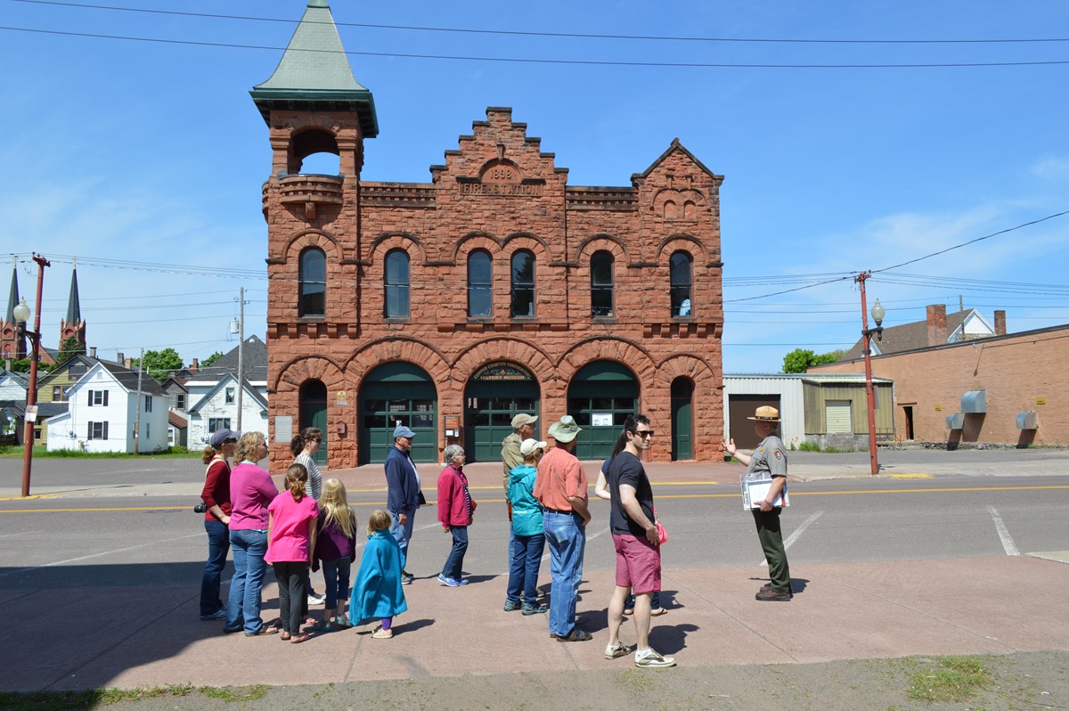 A park ranger speaks to a group of people outside of a two-story sandstone building.