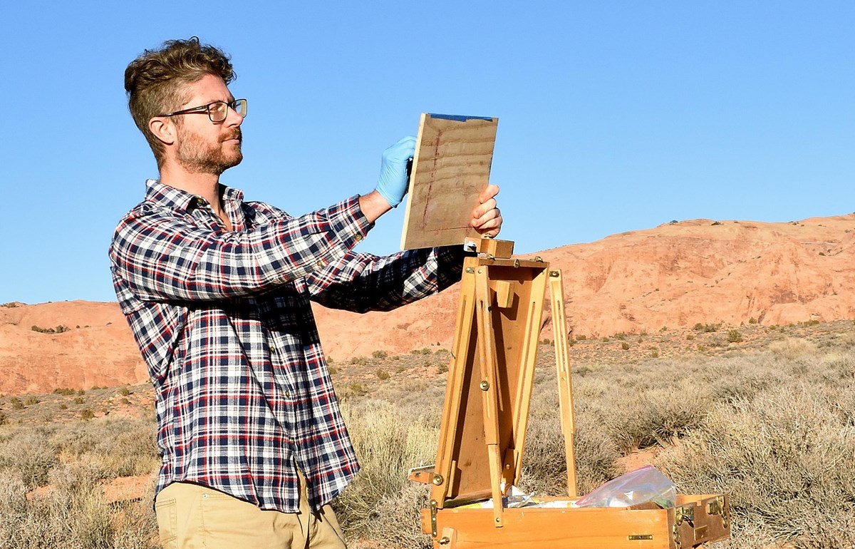 a man holds a wood board next to an easel