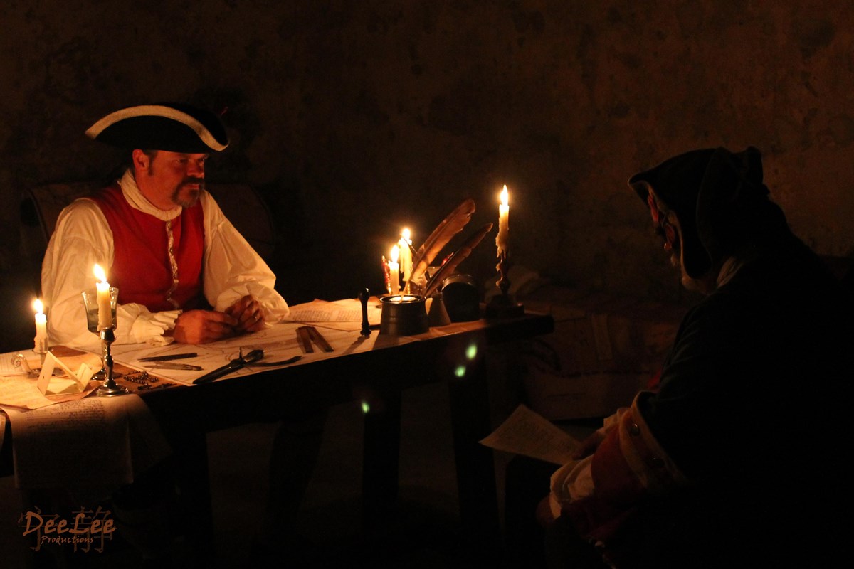 Two colonial spanish soldiers sitting a desk and illuminated by candlelight.