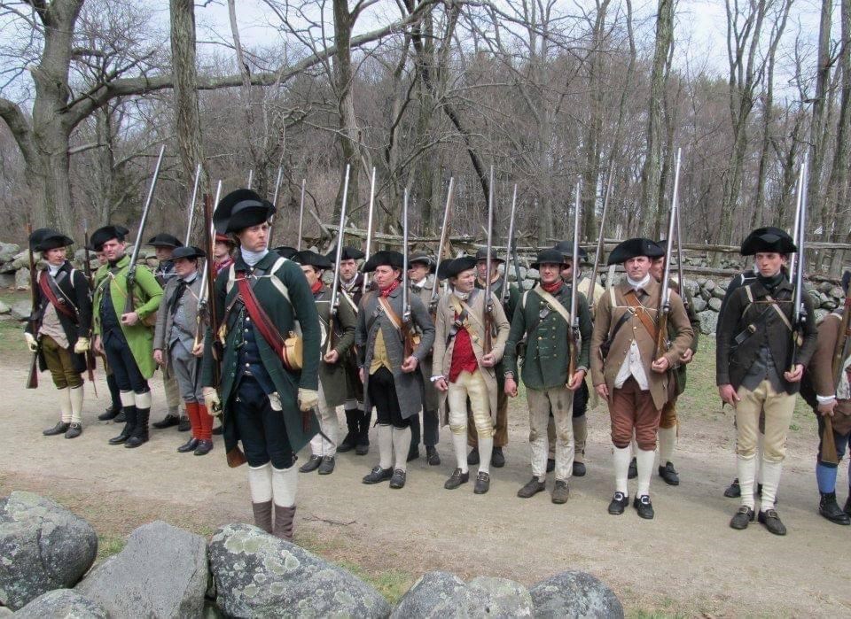 Several Colonial Revolutionary War militiamen stand in a line with muskets.