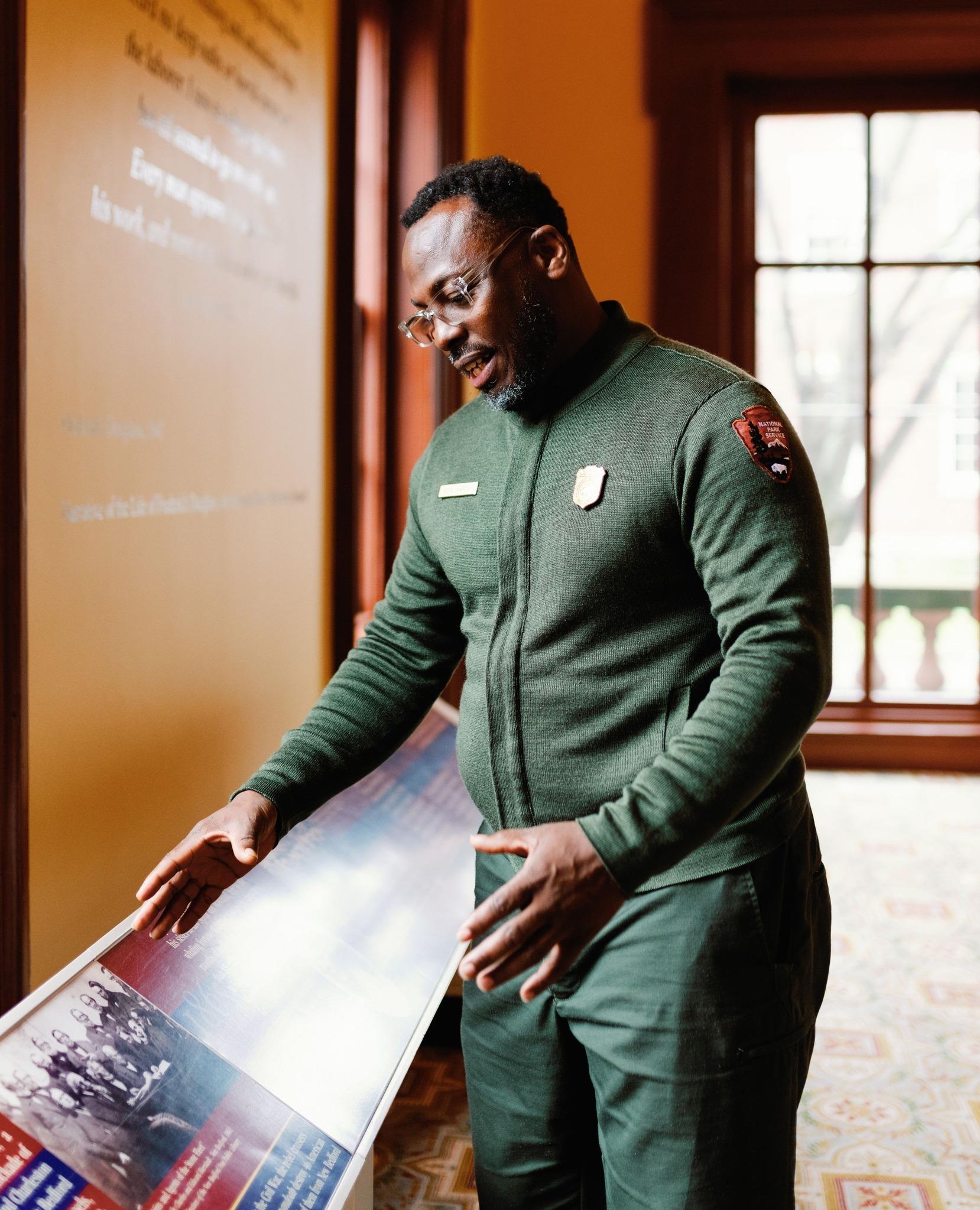Ranger Rufai in uniform gestures toward an indoor exhibit panel inside a historic building
