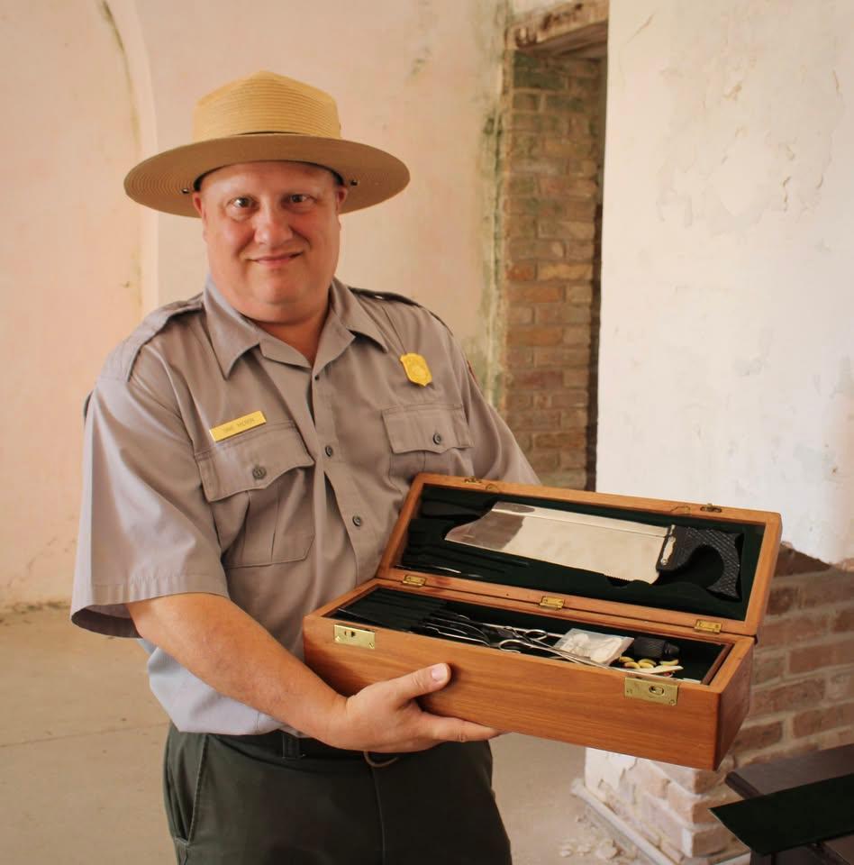A man in grey shirt, green pants and tan hat holds case with tools