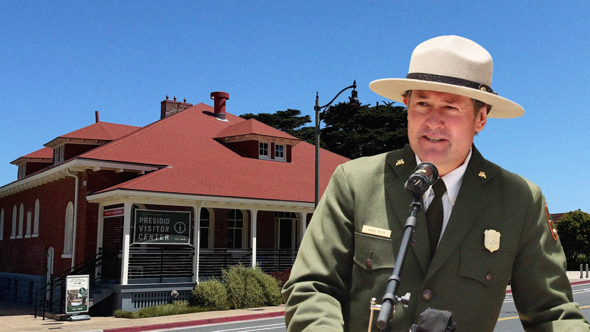 A person in uniform stands in front of a microphone. A building with a red roof is in the background