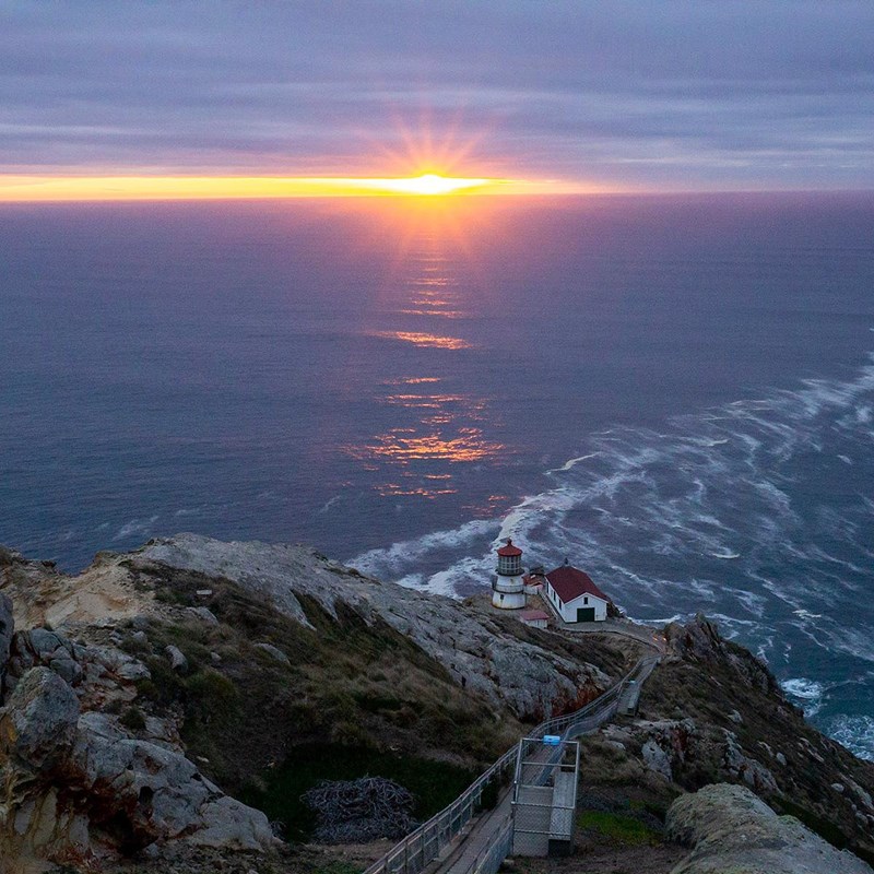 Above a short lighthouse perched on a rocky cliff, the sun sets between clouds & the ocean horizon.