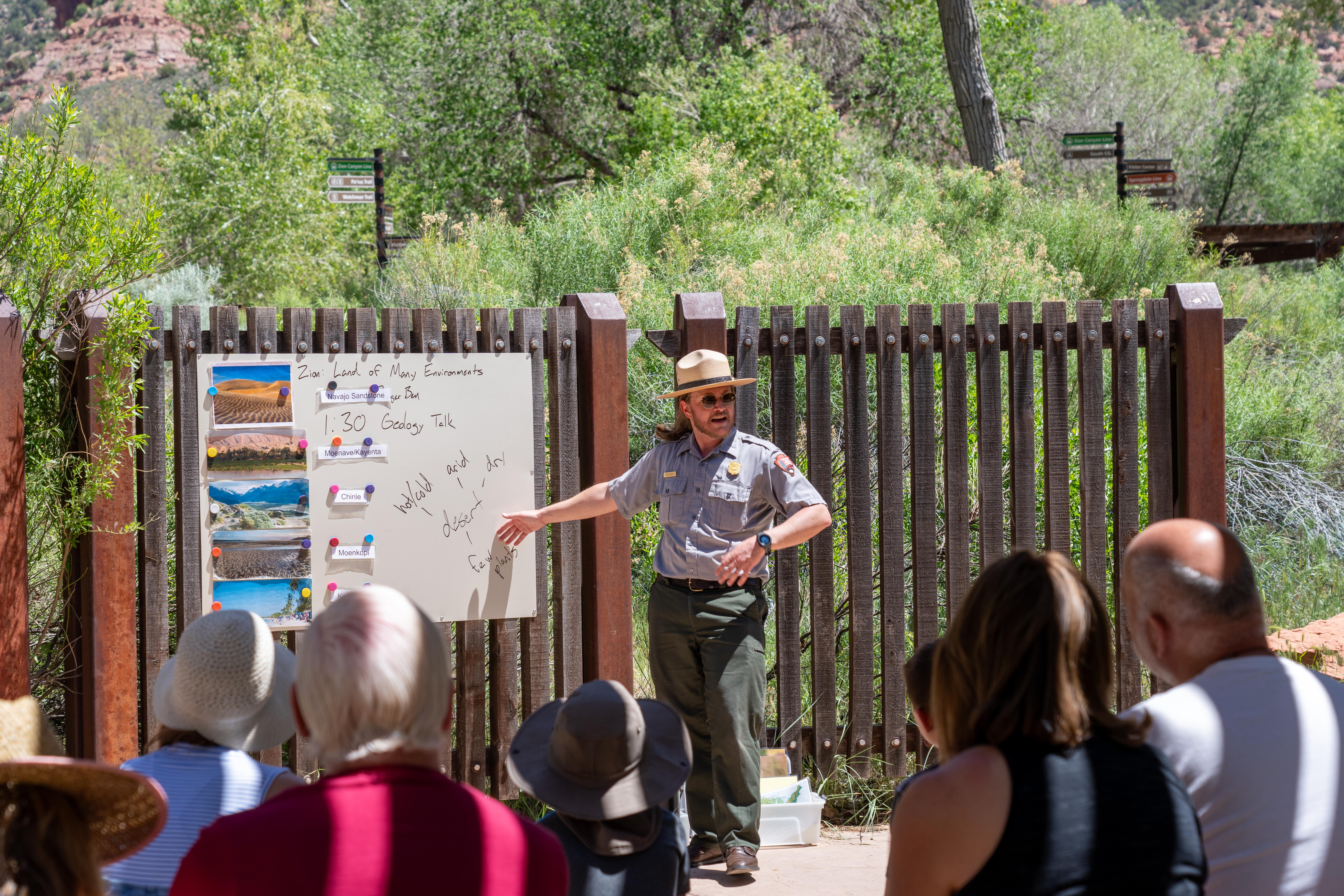 A park ranger in a tan flat hat stands next to a whiteboard speaking to a crowd of seated people.