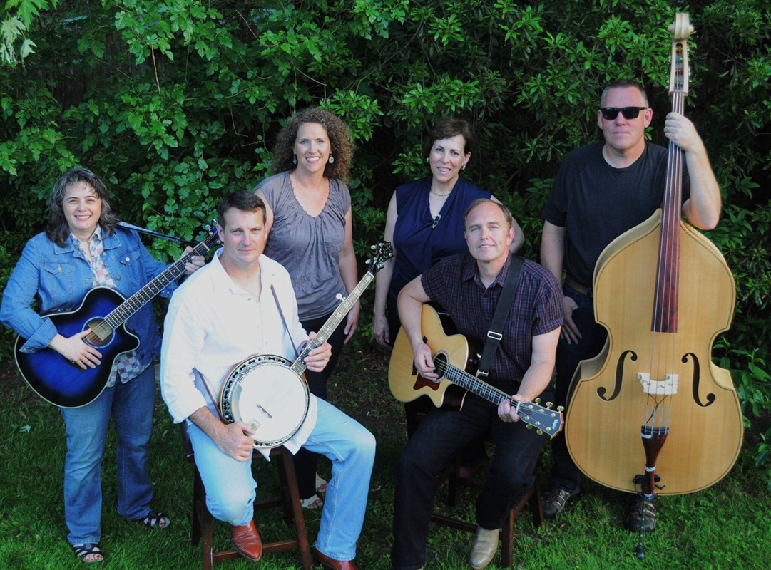 Chisman Creek Band members pose with instruments on green grass against green trees.