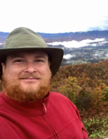 A bearded man in a red jacket and hat smiles in front of a mountain range