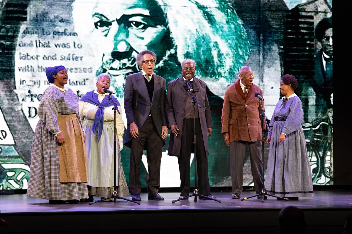 Men and women stand on a stage performing with a large photo of Frederick Douglass behind them.