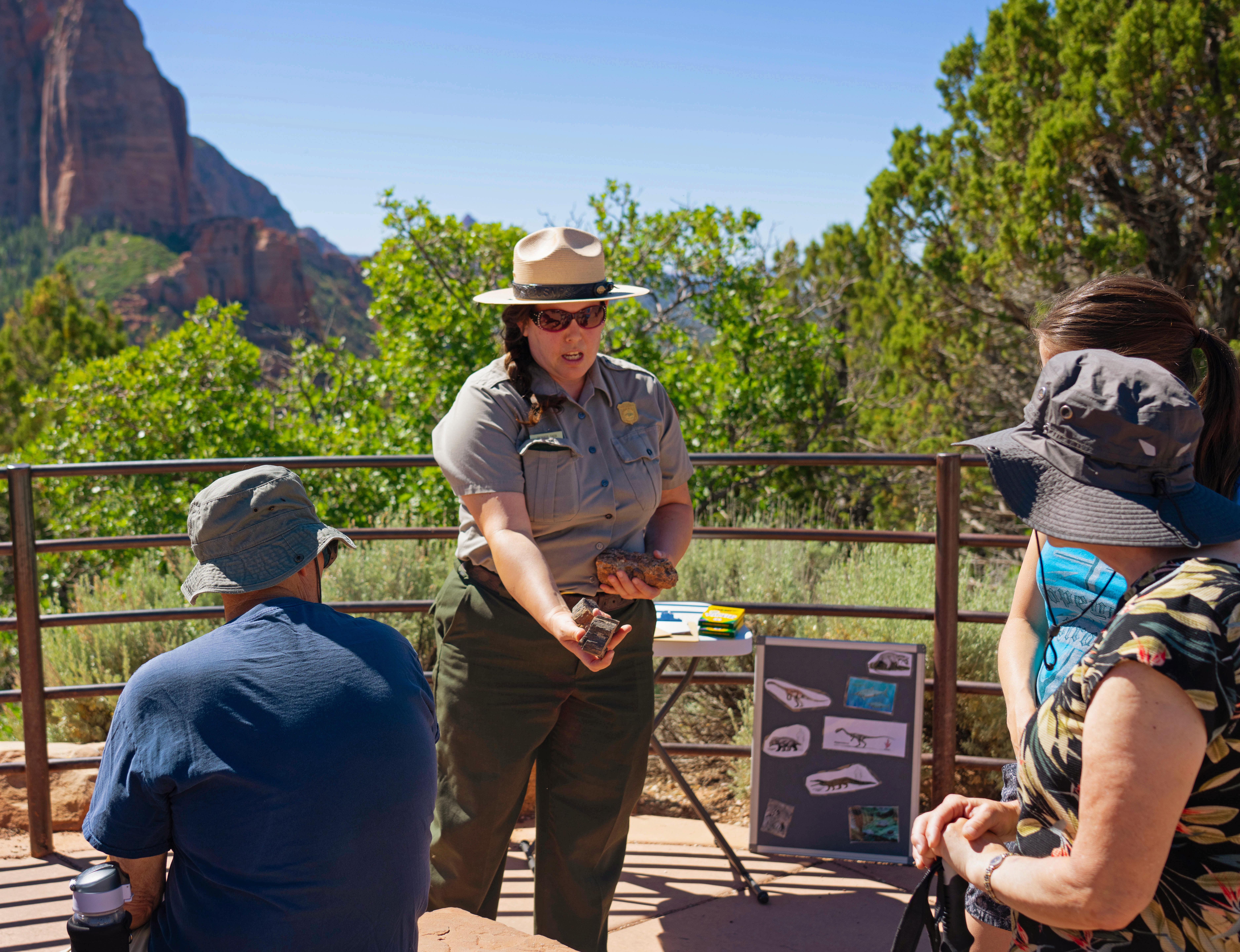 A ranger in dark sunglasses shows a rock to a seated pair of visitors.
