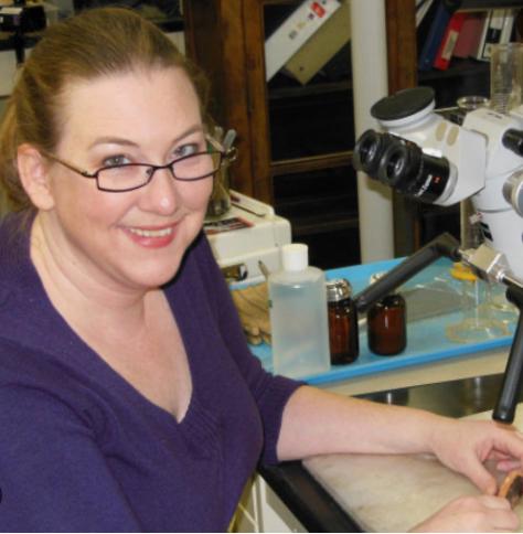 A woman with glasses seated in front of a microscope.