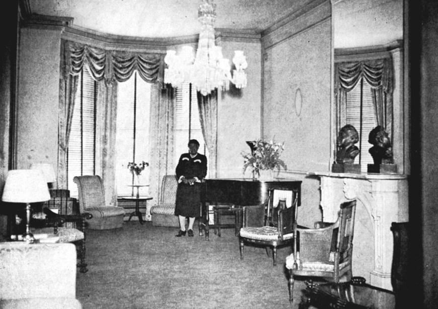 A woman stands in a well-furnished, ornate room in front of large windows, next to a piano.
