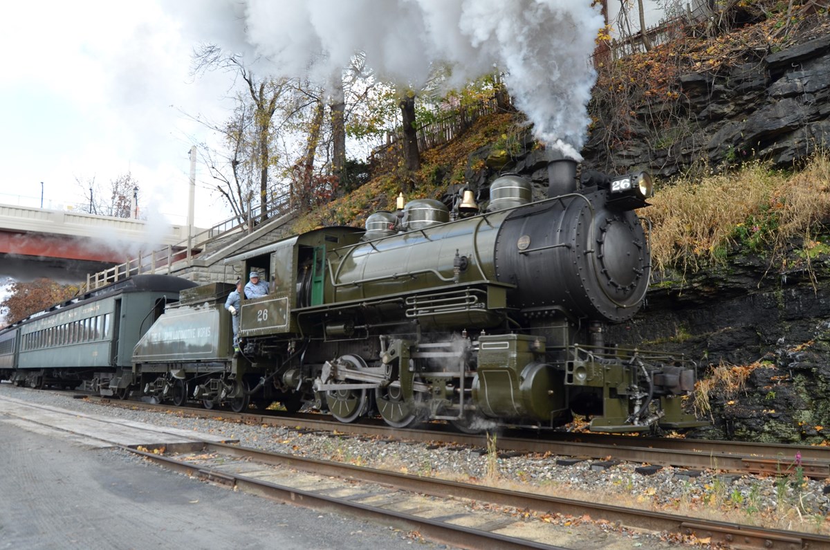 engineer leans out the cab window of a dark green steam locomotive, steam coming from top of engine