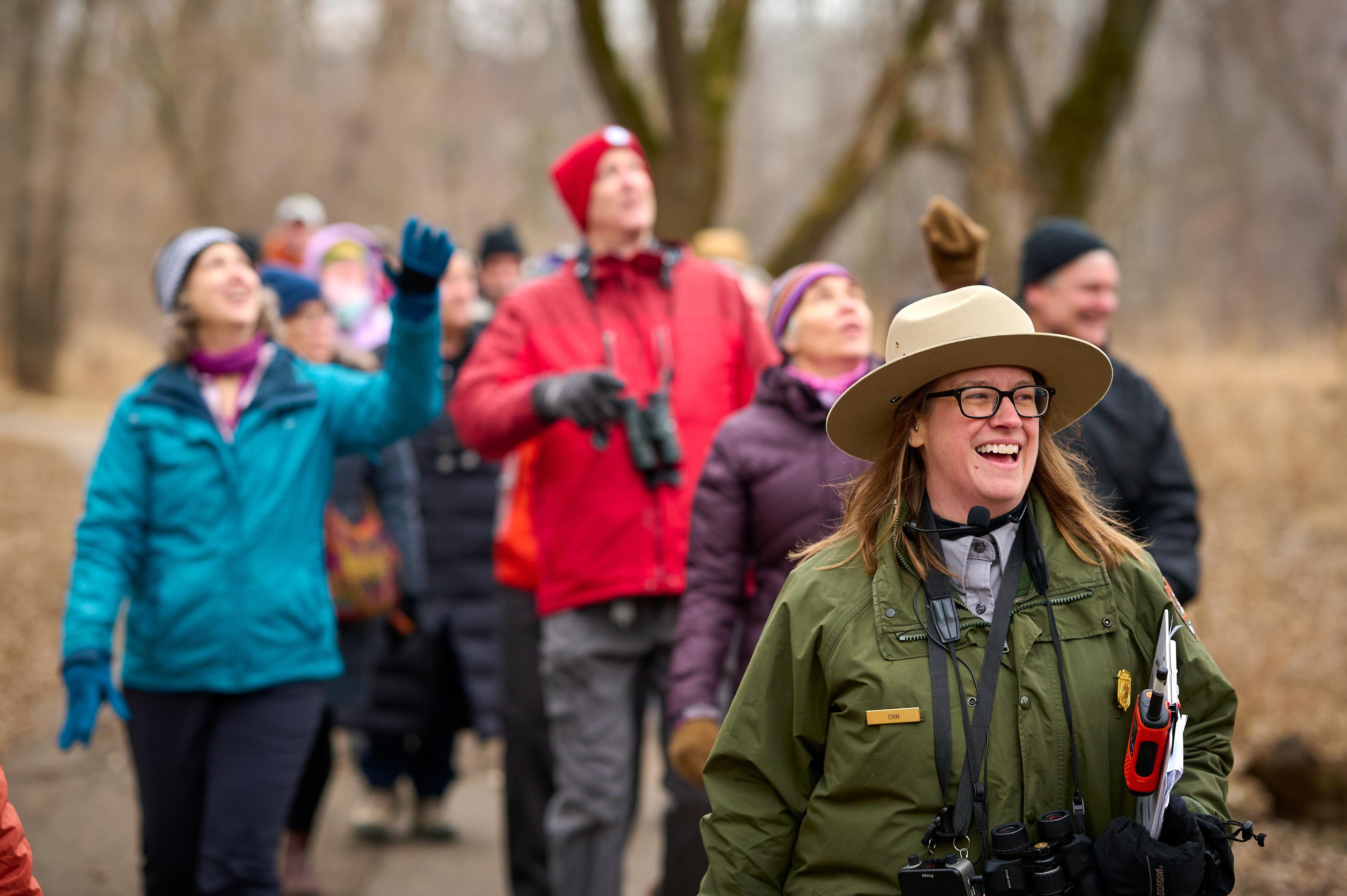 A park ranger smiles as they lead visitors with binoculars on a trail.
