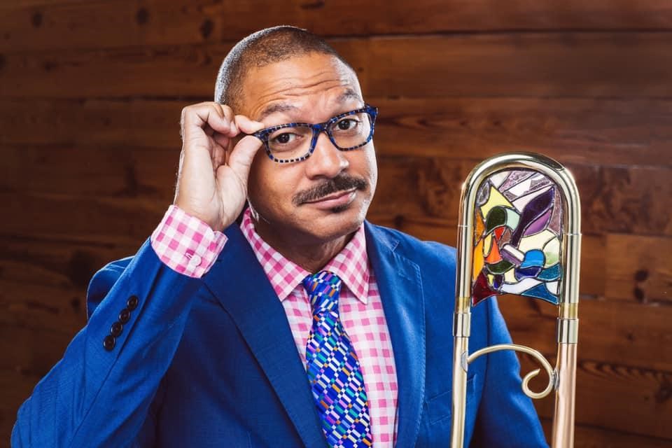 Delfeayo Marsalis portrait wearing bright blue balzer and tie holding trombone and touching glasses.