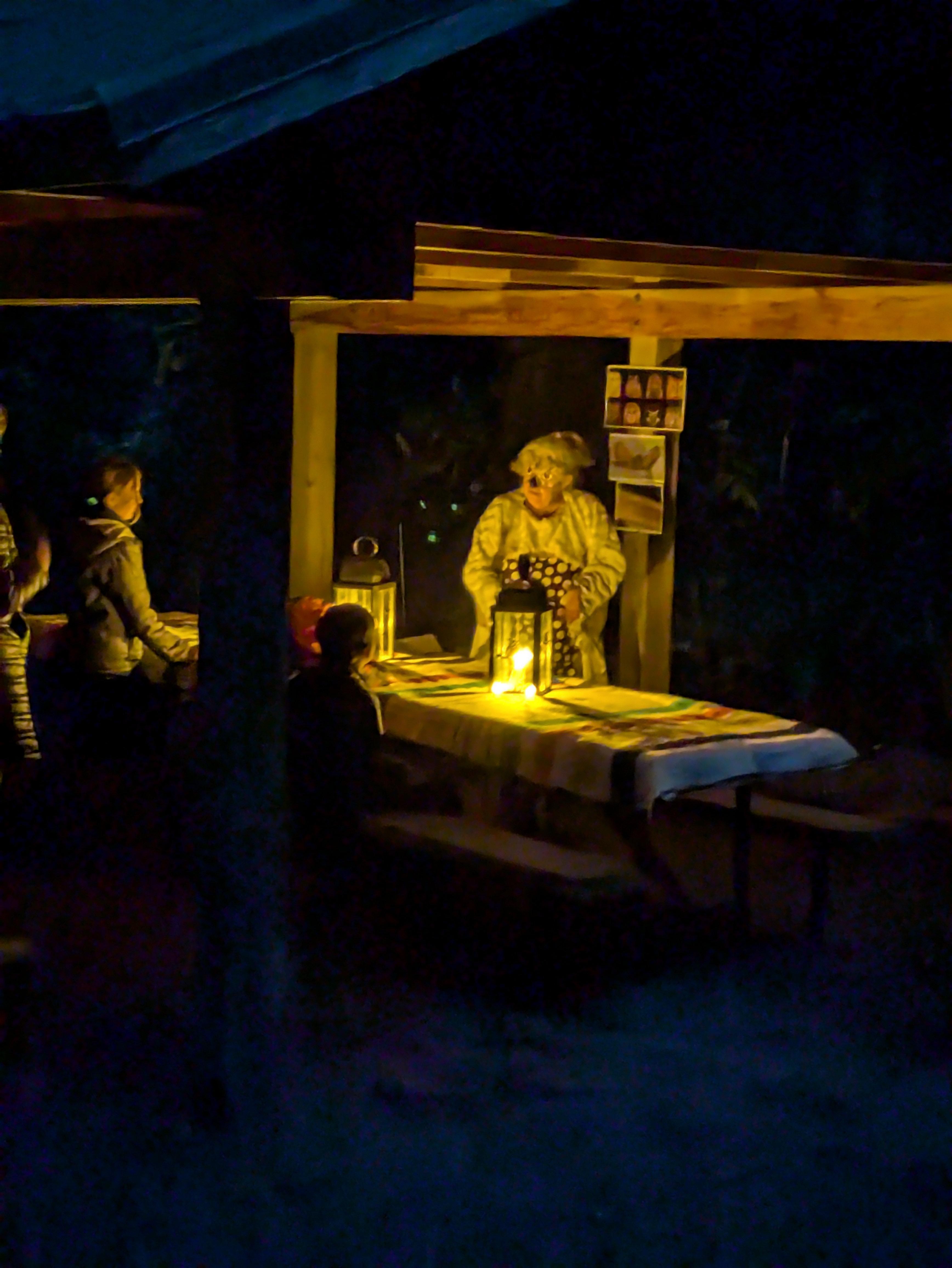 A ranger in an Owl mask talks by lantern light with an audience around a illuminated picnic table