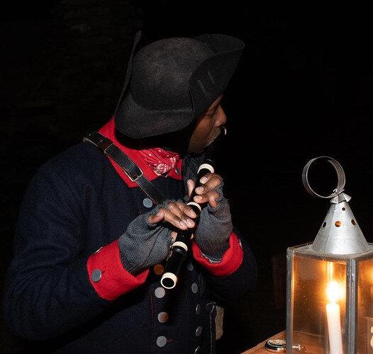 An African American soldier in 18th century uniform plays a fife next to a lantern