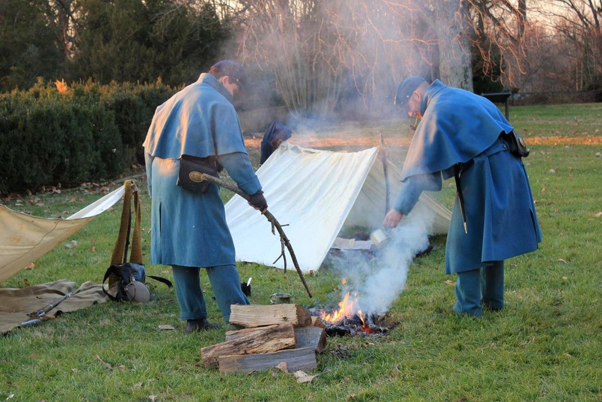Two living historians, U.S. Army Civil War soldiers tend to a fire in front of a canvas tent.