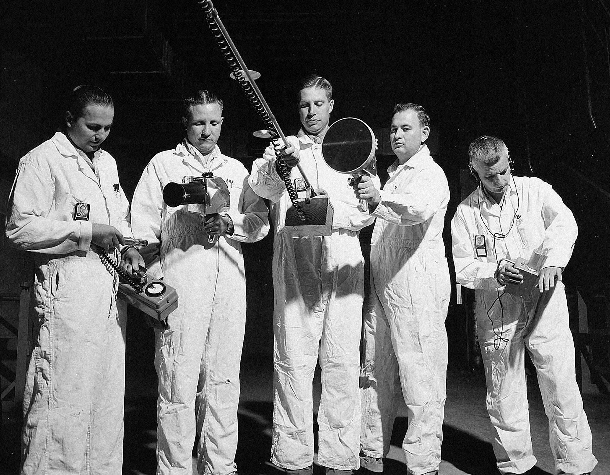 5 men in white coveralls stand in a row holding various handheld devices.