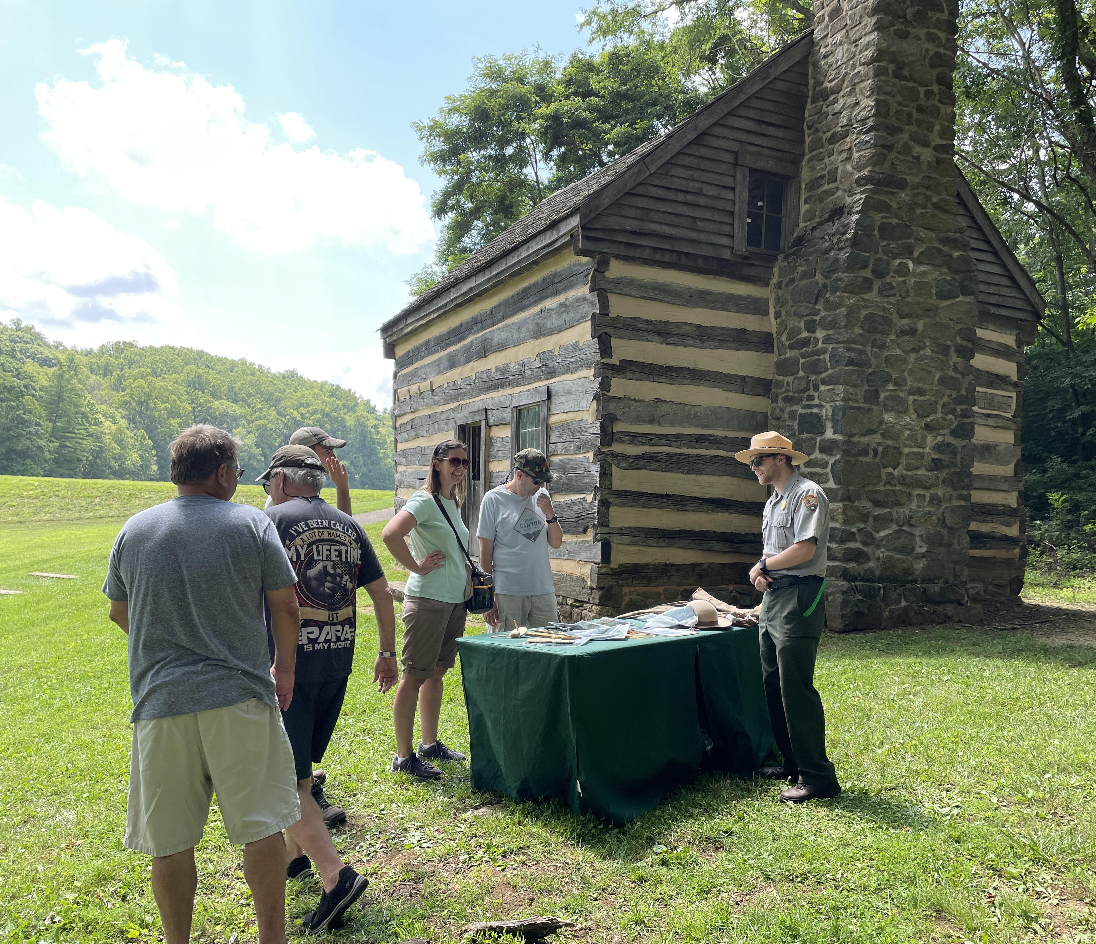 5 adults and a uniformed ranger gather around a table with 2-story cabin in the background