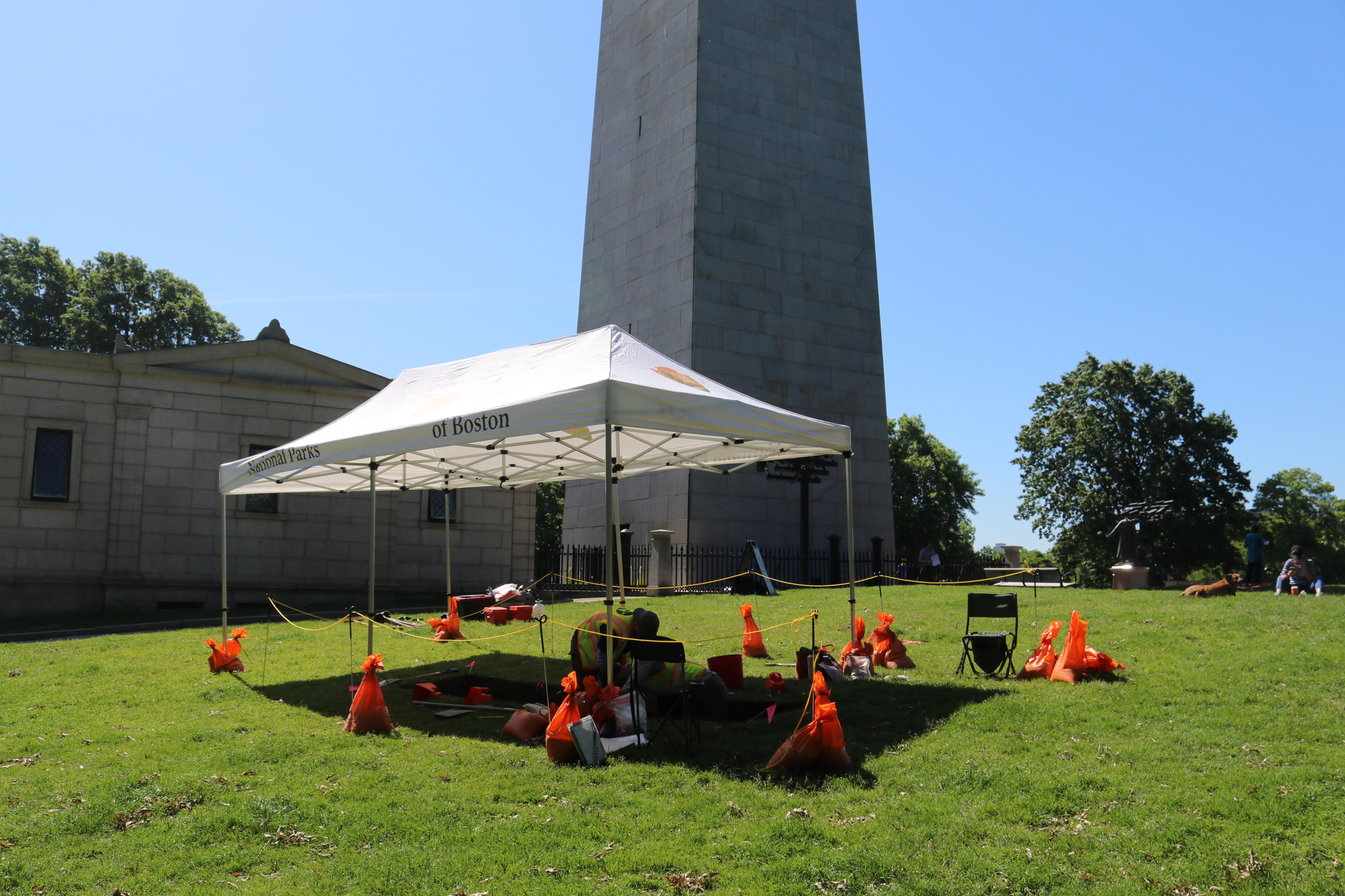 A tent covers workers in vests digging into a grassy lawn site