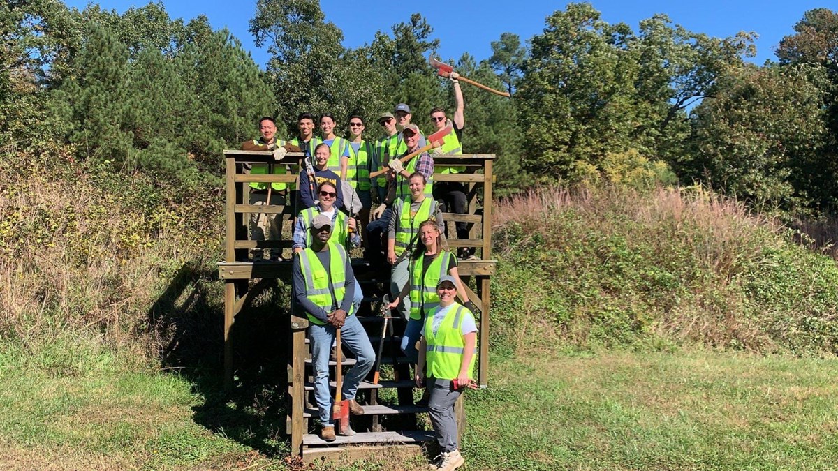 A group of 10 volunteers wearing bright green safety vests stand on a wooden platform.