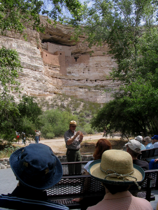 a ranger talking in front of a group of seated visitors in front of a large, adobe cliff dwelling
