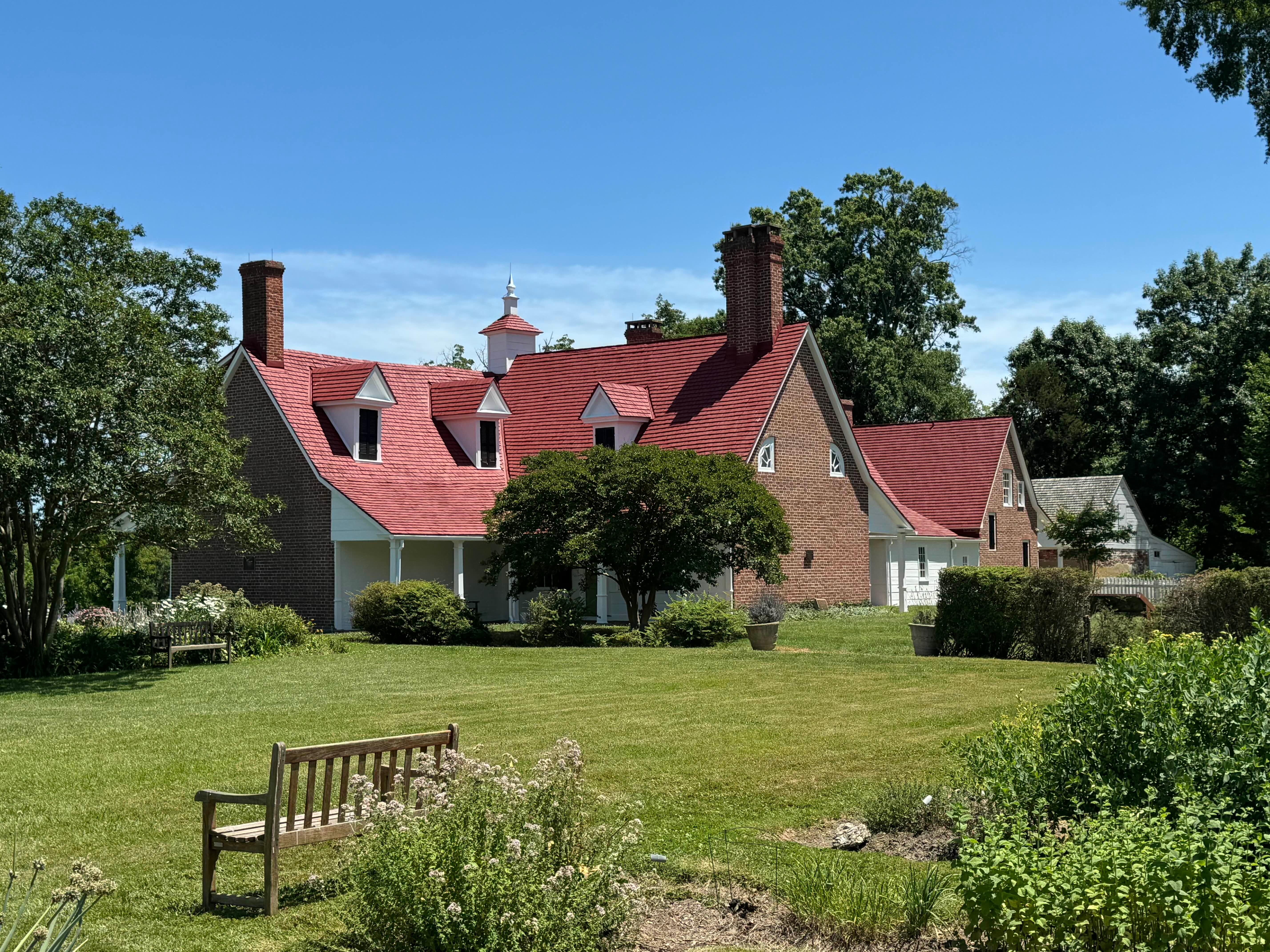 A brick house with a red roof and dormer windows, surrounded by green lawns and trees, under a clear