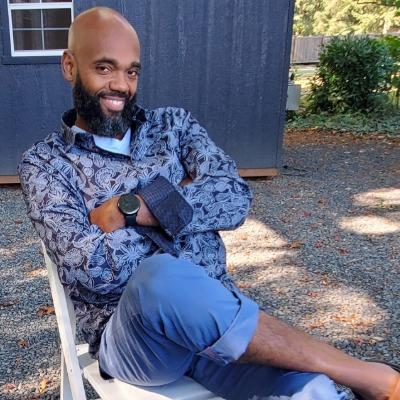 Bearded African American man smiles at camera, ankle crossed over knee and arms crossed.
