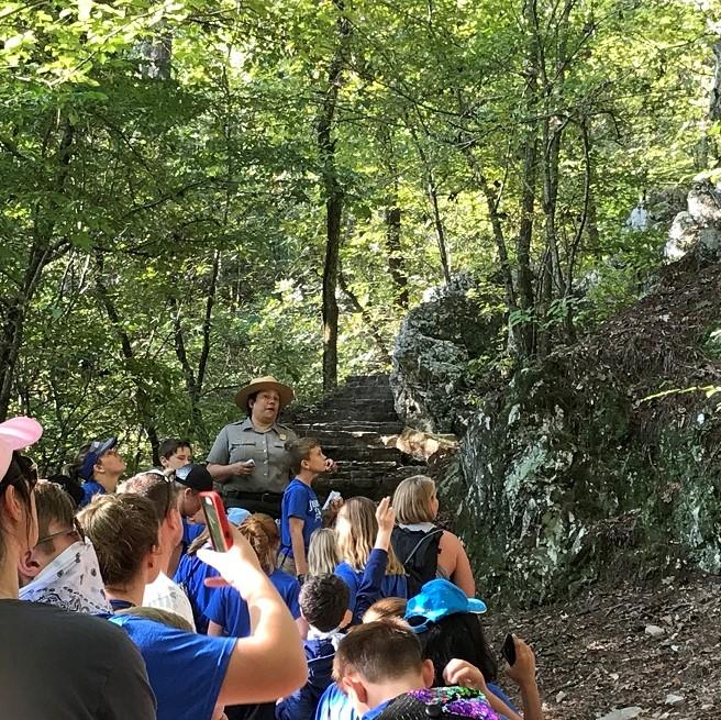 Female ranger stopped on the trail in the green forest talking to a group of young kids