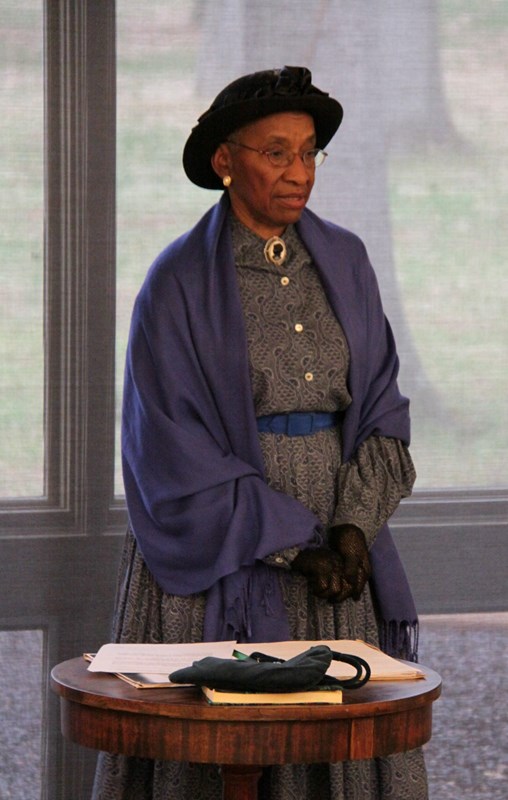 African American woman wearing hat and shawl standing in front of a table