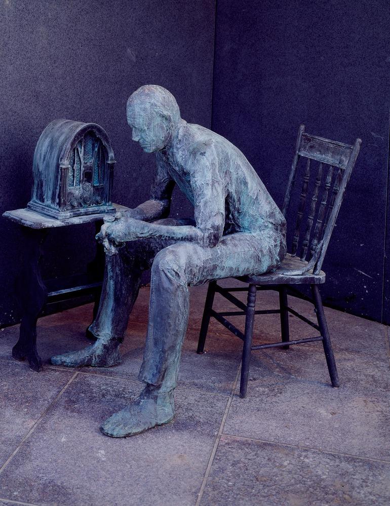 Weathered bronze statue of a man sitting in a chair, listening to a radio