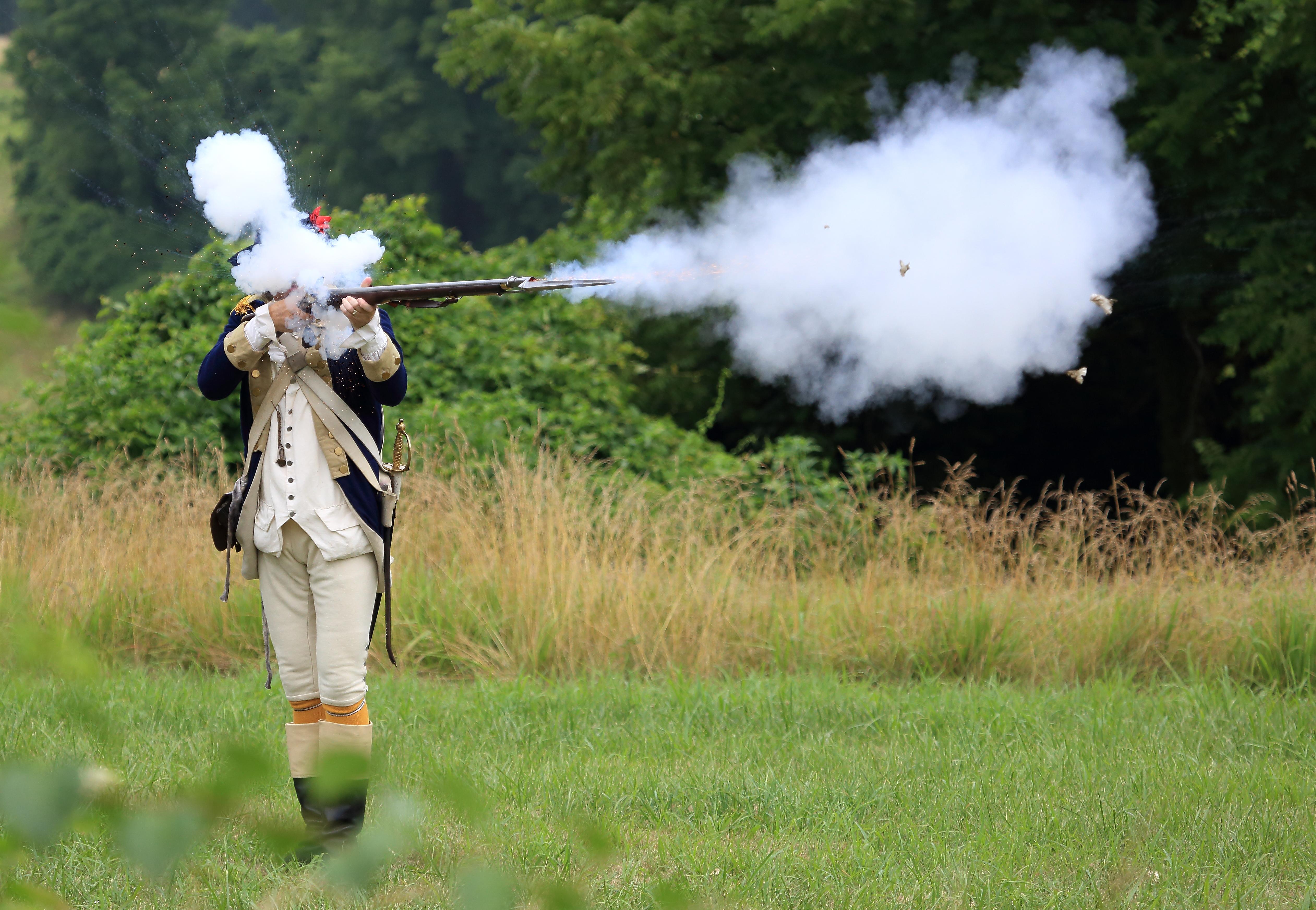 a soldier in 18th century uniform fires a musket and smoke covers his face and erupts out of the gun