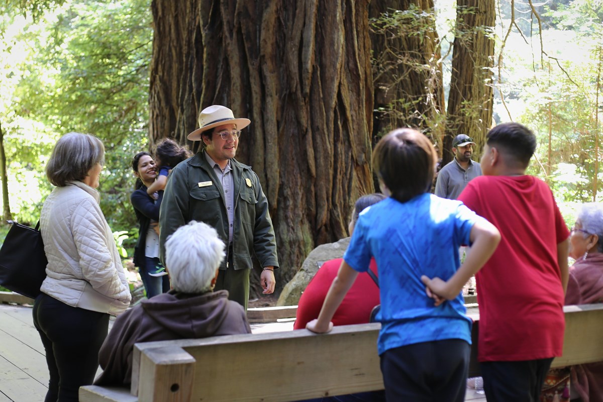standing in front of a massive trunk, a ranger speaks with visitors of many ages
