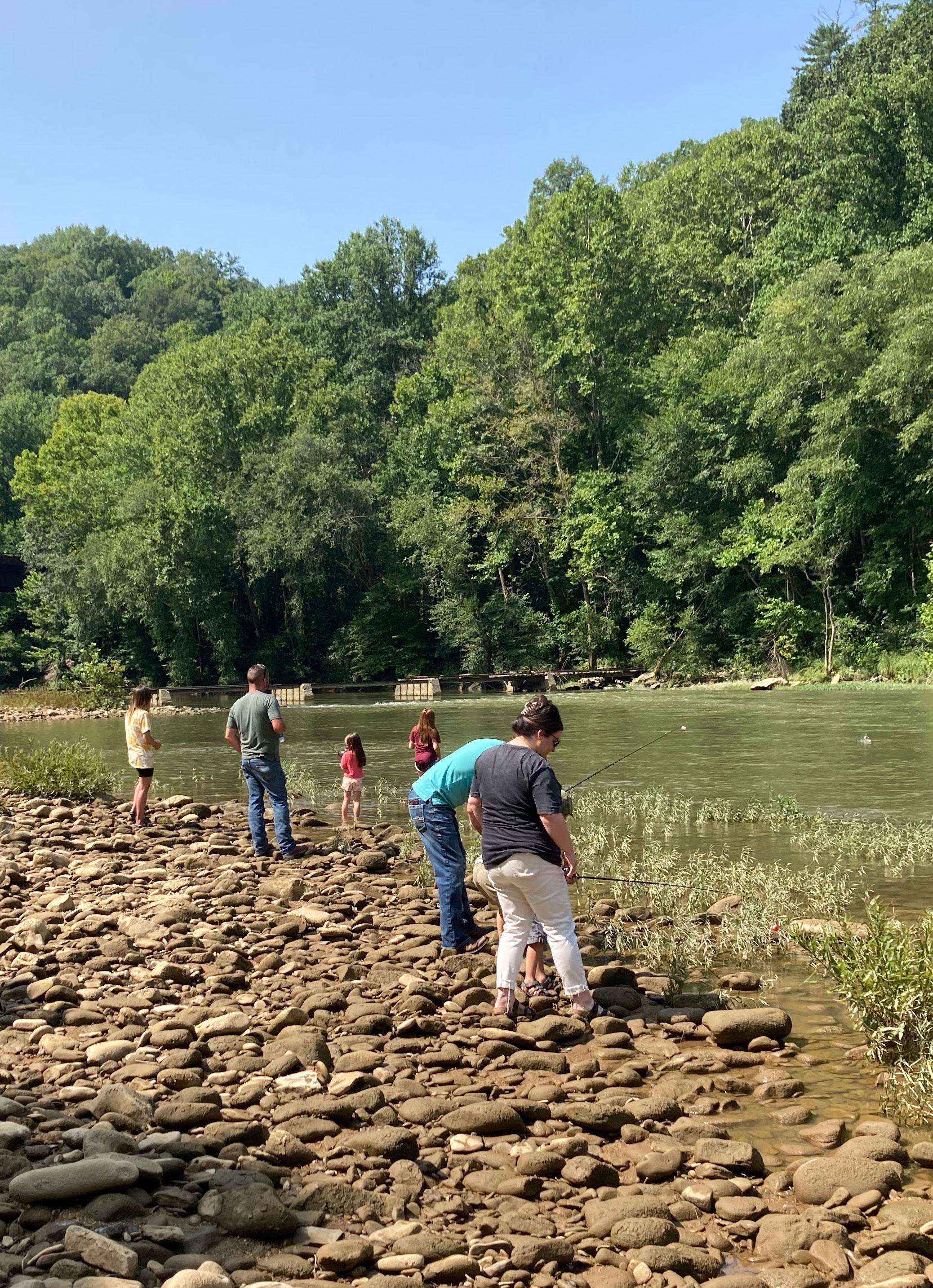 Adults and children stand on a rocky beach, fishing in a forested river stream.